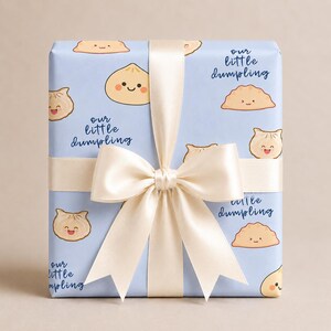 May include: A rectangular gift box wrapped in light blue paper featuring a repeating pattern of cartoon dumplings with smiling faces and the text our little dumpling in dark blue script, tied with a wide, cream-colored satin ribbon bow.