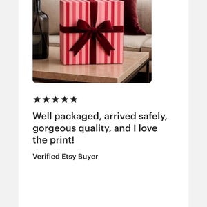 May include: A square gift box featuring a vertical striped pattern in alternating shades of light and dark pink. The box is wrapped with a dark red velvet ribbon tied into a centred bow on the front face, creating a decorative presentation.