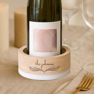 May include: A wooden wine bottle holder with a white base, holding a wine bottle with a pink label. The holder is engraved with "the johnsons" and a leaf design. A gold fork and wine glasses are in the background.