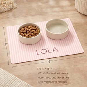 Puede incluir: Una alfombrilla para mascotas a rayas rosas y blancas con el nombre "LOLA" impreso. Dos cuencos de cer&aacute;mica blanca se encuentran sobre la alfombrilla, uno lleno de comida para mascotas y el otro con agua. La alfombrilla mide 30 cm por 46 cm.