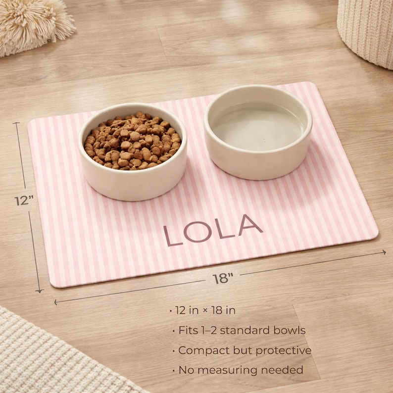 Puede incluir: Una alfombrilla para mascotas a rayas rosas y blancas con el nombre "LOLA" impreso. Dos cuencos de cer&aacute;mica blanca se encuentran sobre la alfombrilla, uno lleno de comida para mascotas y el otro con agua. La alfombrilla mide 30 cm por 46 cm.