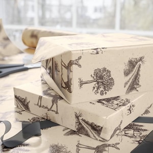 May include: Two gift boxes wrapped in cream-colored paper with a black and white forest scene print. The boxes are stacked, with one partially unwrapped. A roll of paper, scissors, and a dark gray ribbon are also visible.