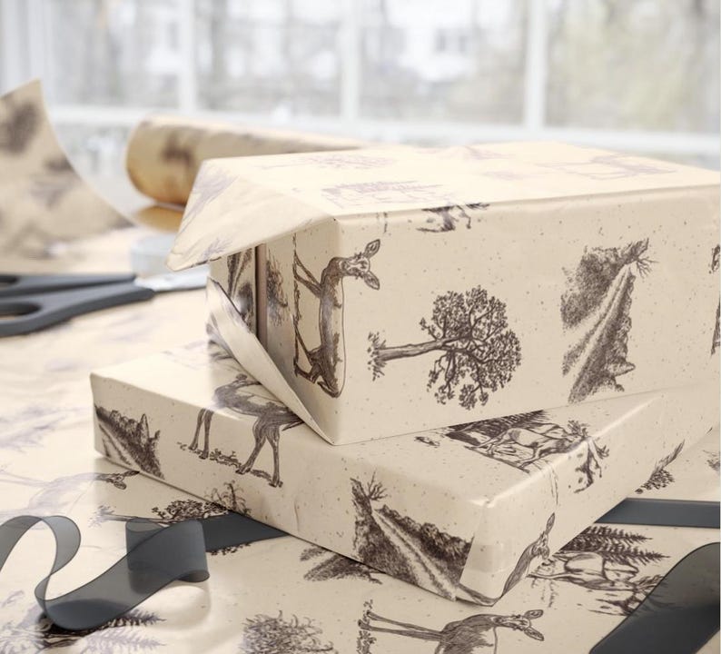 May include: Two gift boxes wrapped in cream-colored paper with a black and white forest scene print. The boxes are stacked, with one partially unwrapped. A roll of paper, scissors, and a dark gray ribbon are also visible.