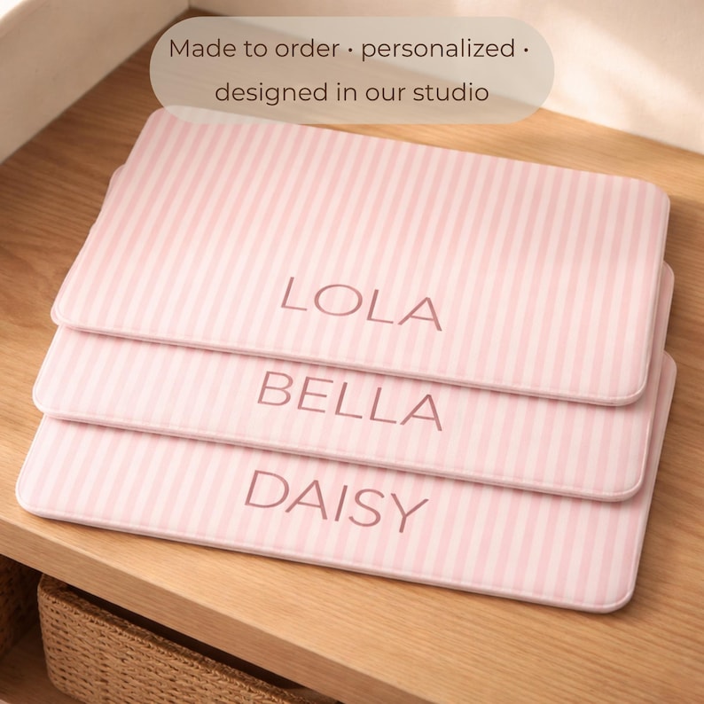 Puede incluir: Tres alfombras rosas a rayas con los nombres Lola, Bella y Daisy. Las alfombras est&aacute;n personalizadas y dise&ntilde;adas en un estudio. Est&aacute;n apiladas en un estante de madera.