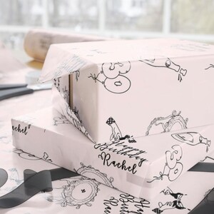 K&ouml;nnte beinhalten: Zwei rechteckige Geschenkboxen, eingewickelt in hellrosa Papier mit einem sich wiederholenden schwarzen Linienmuster aus Luftballons mit der Zahl 30, Champagnerflaschen und dem Text Happy Birthday Rachel in einer kursiven Schriftart.
