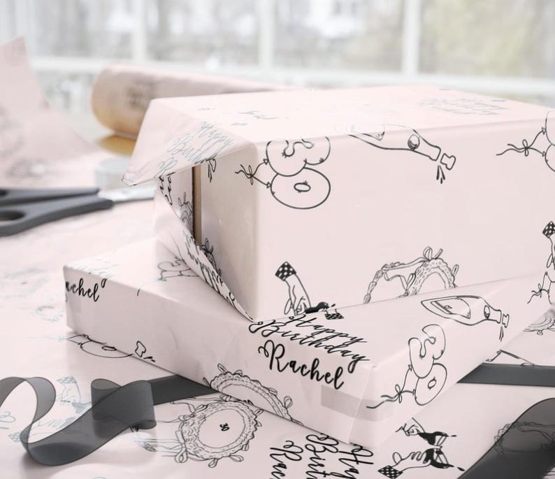 K&ouml;nnte beinhalten: Zwei rechteckige Geschenkboxen, eingewickelt in hellrosa Papier mit einem sich wiederholenden schwarzen Linienmuster aus Luftballons mit der Zahl 30, Champagnerflaschen und dem Text Happy Birthday Rachel in einer kursiven Schriftart.
