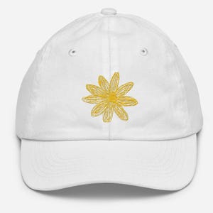 Może przedstawiać: Biała czapka z daszkiem z haftowaną żółtą stokrotką z przodu.
