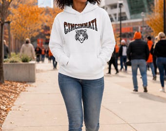 May include: A white hooded sweatshirt featuring a front kangaroo pocket and a graphic print on the chest. The graphic displays the word CINCINNATI in a stylised font with a tiger face illustration positioned directly below the text.