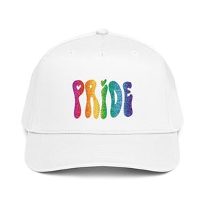 Op de afbeelding: Een witte baseballcap met het woord "PRIDE" in regenboogglitterletters. De letters zijn in een golvend, bubbelachtig lettertype, met een klein hartje ter vervanging van de letter "I". De cap heeft een gebogen klep en een knoop aan de bovenkant.