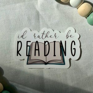 May include: White sticker with black text that reads "I'd rather be reading" with a cartoon illustration of an open book with teal accents.