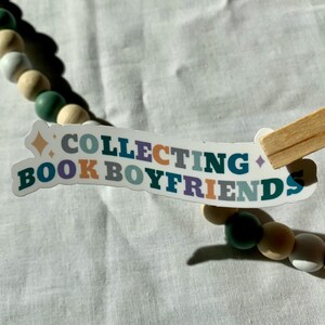 May include: A white sticker with the text "COLLECTING BOOK BOYFRIENDS" in colorful letters. The sticker is slightly curved and has a small diamond shape in the top left corner.