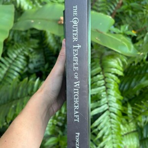 Pode incluir: Lombo de livro escuro com o t&iacute;tulo "The Outer Temple of Witchcraft" e o nome do autor, Penczak, em texto branco. O lombo tem um gradiente verde e um s&iacute;mbolo triangular no topo. O livro &eacute; segurado contra um fundo de folhagem verde.