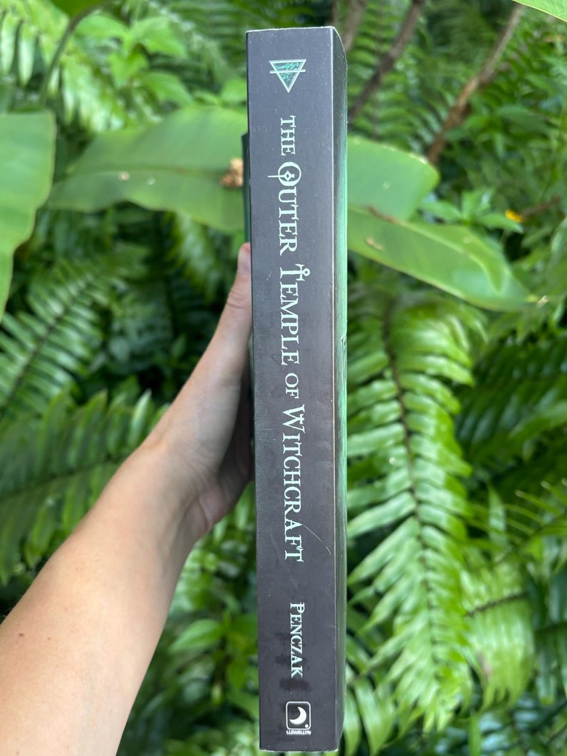 Pode incluir: Lombo de livro escuro com o t&iacute;tulo "The Outer Temple of Witchcraft" e o nome do autor, Penczak, em texto branco. O lombo tem um gradiente verde e um s&iacute;mbolo triangular no topo. O livro &eacute; segurado contra um fundo de folhagem verde.
