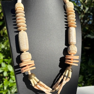 May include: A long necklace made of carved bone beads in shades of cream and peach. The necklace features a variety of bead shapes, including cylindrical, round, and flat. The necklace is accented with small gold-toned beads.