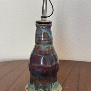 May include: A handcrafted ceramic oil bottle with a stainless steel spout and black cap. The bottle features a unique glaze in shades of blue, purple, and green. The base is a light tan color, and the bottle sits on a wooden surface.