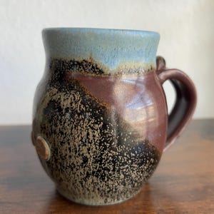 May include: Handmade ceramic mug with a unique speckled design. The mug features a dark brown handle and a rim with a blue-green glaze. The body of the mug has a speckled pattern in shades of brown, black, and tan. A small, round, decorative element is on the side.