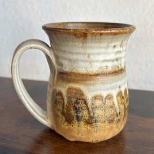 May include: A ceramic mug featuring a flared rim and a curved handle. The upper portion has a smooth, off-white glaze, while the lower section displays a textured, mottled brown and tan drip pattern that extends toward the base of the vessel.