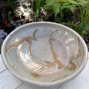 May include: A ceramic bowl with a light gray glaze and brown and green streaks. The bowl is sitting on a white wooden surface.