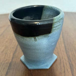 May include: A handmade ceramic tumbler with a unique design. The top rim is black, transitioning to a light blue body with a hexagonal base. The interior is black.