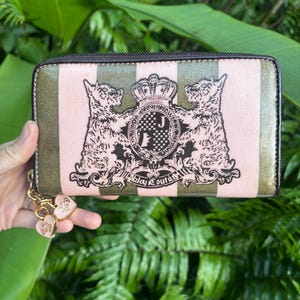 May include: A rectangular wallet with a pink and olive green striped pattern. The wallet features a crest with two dog illustrations and the text "Juicy Couture". A gold chain with heart-shaped charms is attached.