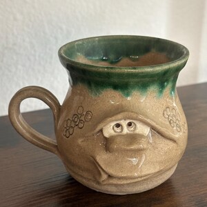 May include: A handmade ceramic mug with a whimsical face design. The mug features a tan body with a green rim and handle. The face has two small eyes and a smiling mouth. Decorative floral patterns are embossed on the sides.