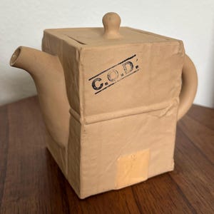 May include: A tan ceramic teapot with a square body and a lid with a small knob. The teapot has a handle and a spout. The text "C.O.D." is stamped on the front. The teapot is designed to look like a cardboard box.