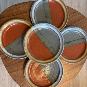 May include: A set of five ceramic plates with a unique design. Each plate features a diagonal split with a burnt orange and gray-green color scheme. The plates have a beige rim and are arranged on a wooden surface.