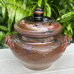 May include: A handcrafted ceramic lidded pot with a rich, earthy color palette. The pot features a rounded body with two handles and a lid topped with a small knob. The glaze has a mix of brown, black, and reddish-orange hues, creating a unique, rustic appearance.