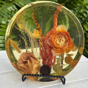 May include: A round, clear resin art piece with dried flowers and foliage encased within. The arrangement features orange and tan roses, red stems, and various dried plants. The piece is displayed on a black metal stand.