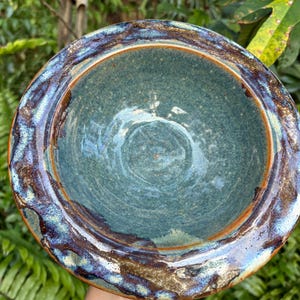 May include: A handmade ceramic bowl with a teal interior and a textured, multi-toned rim. The rim features shades of blue, brown, and white, creating a unique, organic design. The bowl's interior has a glossy finish, reflecting light. The bowl is held against a backdrop of green foliage.