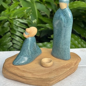 May include: A handcrafted nativity scene featuring three ceramic figures in teal robes and tan accents, set on a natural wood base. The figures include two standing and one kneeling, with a small bowl. The scene evokes a sense of peace and spirituality.