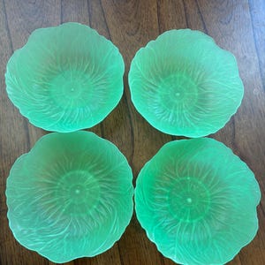 May include: Four green glass plates with a ruffled edge and a textured, swirling pattern. The plates are arranged on a dark wooden surface. The plates have a unique, organic design.