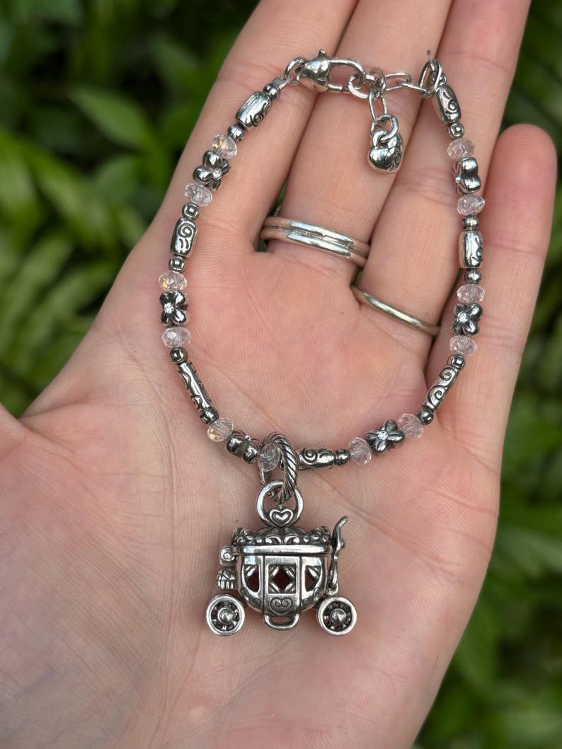 May include: Silver bracelet featuring a carriage charm. The bracelet has a mix of clear and pink beads, with decorative silver beads. The carriage charm has intricate details and a red center. The bracelet is secured with a lobster clasp.
