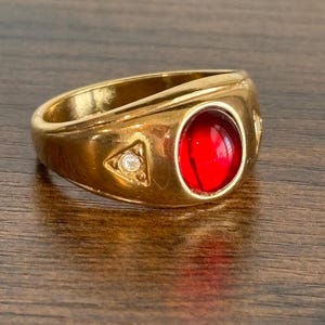May include: A gold-toned ring featuring a central oval-shaped red cabochon stone. The band includes a small triangular accent on the side containing a single clear, round-cut stone set within the metal surface, creating a polished appearance.