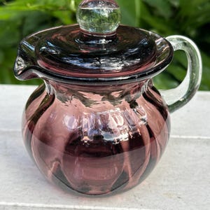 May include: A small, purple glass creamer with a matching lid and a clear glass handle. The creamer has a rounded body with vertical ridges and a spout for pouring. The lid has a small, round knob.