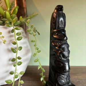 May include: A black carved totem pole featuring a bird at the top, with other animal figures carved below. The totem pole is displayed on a wooden surface, with a white pot of greenery in the background.