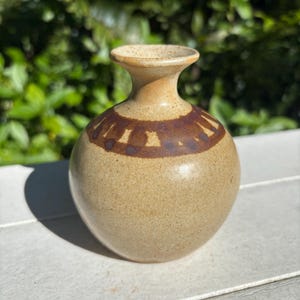 May include: A small, light brown ceramic vase with a brown band around the middle. The vase has a speckled texture and a slightly flared opening.