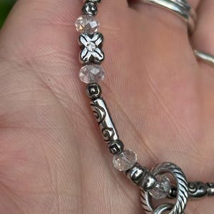 May include: Close-up of a silver-toned necklace with clear crystal beads and decorative metal accents. The necklace features a floral-shaped centerpiece and a twisted metal clasp. The design includes a mix of smooth and textured elements.