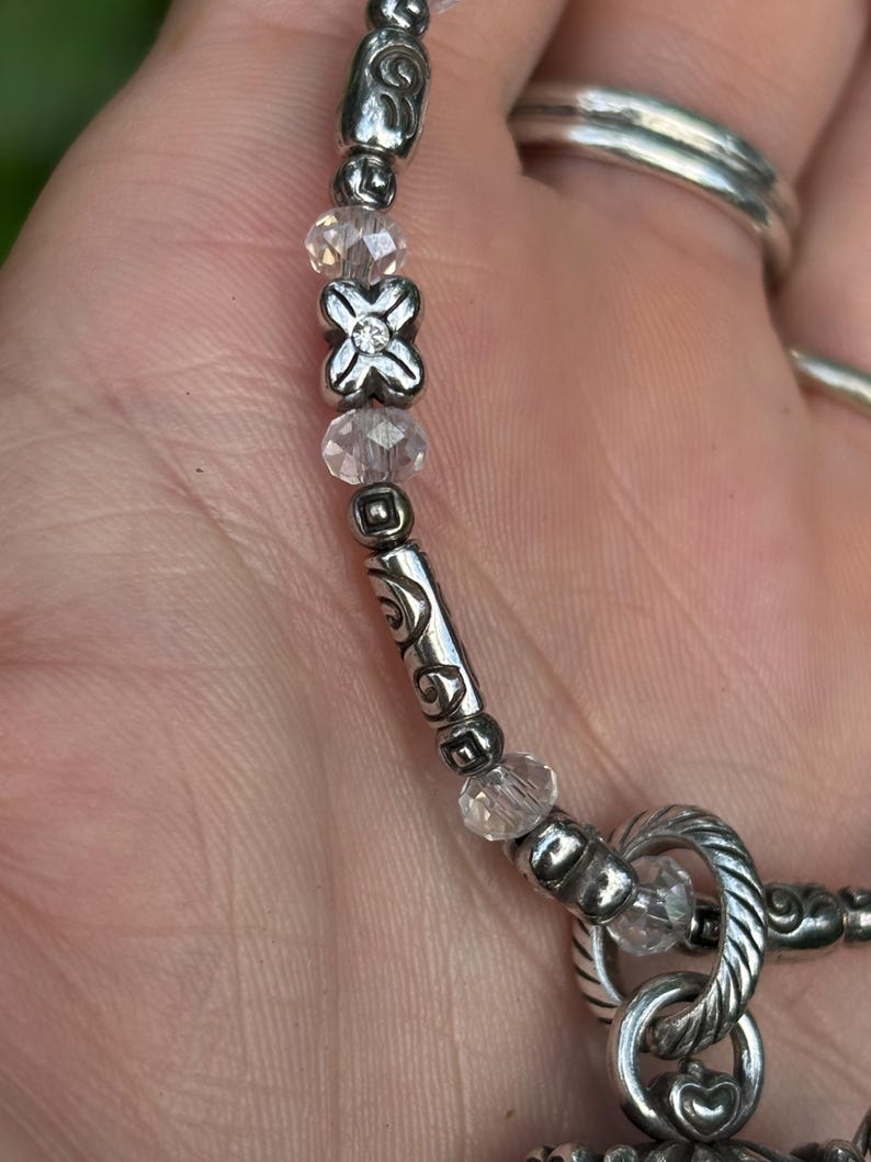 May include: Close-up of a silver-toned necklace with clear crystal beads and decorative metal accents. The necklace features a floral-shaped centerpiece and a twisted metal clasp. The design includes a mix of smooth and textured elements.