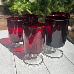 May include: Set of five red glass stemmed drinking glasses with clear glass bases. The glasses are on a white wooden surface.