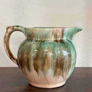 May include: A ceramic pitcher with a rounded body, a handle, and a spout. The pitcher is glazed with a blend of green, brown, and cream colors. The handle and spout are a lighter shade of brown. The pitcher is sitting on a dark wooden surface.