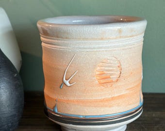 May include: A handmade ceramic mug with a unique design. The mug features a light beige rim and a body with orange and tan hues. It has a textured surface with a white abstract design and a sun-like imprint. The base has black and blue stripes.