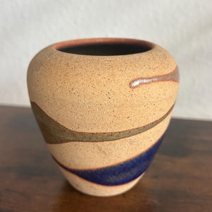 May include: A small, rounded ceramic vase with a speckled beige, matte-textured surface. The vase features horizontal, flowing bands of dark blue and muted olive green glaze that wrap around the body, contrasting with the natural clay finish.