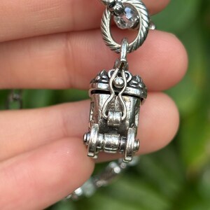 May include: A detailed silver-toned pendant necklace featuring a small, ornate carriage charm. The charm has intricate detailing, including wheels and a hinged door. The necklace is held by a hand against a blurred green background.