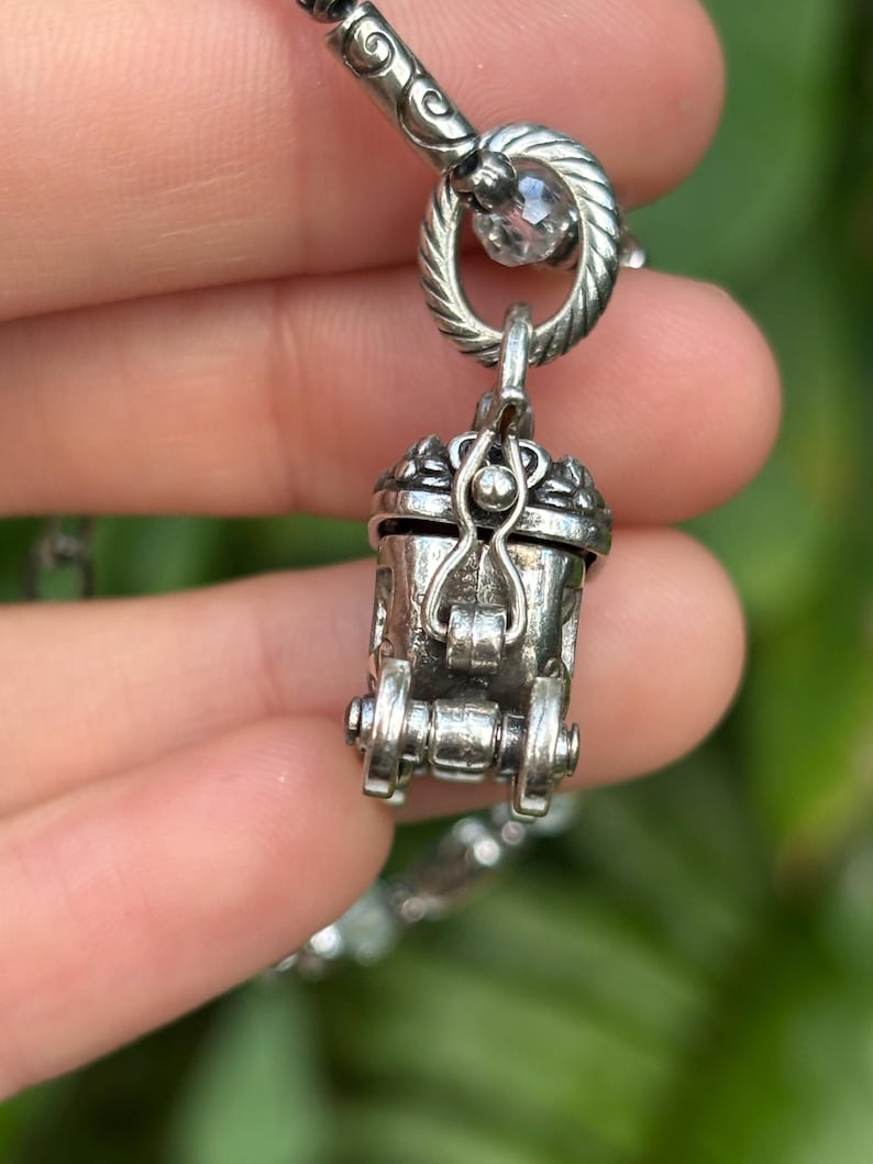 May include: A detailed silver-toned pendant necklace featuring a small, ornate carriage charm. The charm has intricate detailing, including wheels and a hinged door. The necklace is held by a hand against a blurred green background.