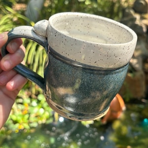 May include: A handmade ceramic mug with a speckled white glaze and a dark blue glaze. The mug has a rounded body and a handle.