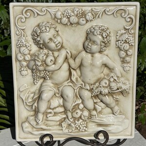 May include: Off-white ceramic relief sculpture featuring two cherubic figures holding fruit and grapes. The square plaque has a decorative border with more fruit and is mounted on a wrought iron stand. The sculpture is a classical decorative piece.