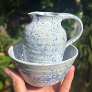 May include: A ceramic pitcher and bowl set. The pitcher has a handle and a spout. Both pieces are white with blue splatter paint. The bowl is round and sits beneath the pitcher. The set is held in a hand.