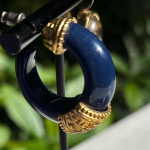 May include: A pair of blue and gold hoop earrings. The hoops are made of a dark blue material and have a gold trim. The earrings are on a display stand.