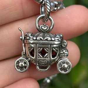 May include: A silver-colored carriage pendant with intricate detailing, including heart-shaped accents and decorative wheels. The carriage windows reveal a red interior. The pendant is suspended from a beaded chain, creating a whimsical accessory.